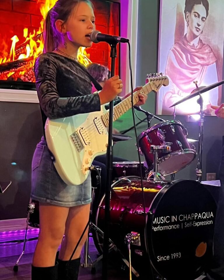A girl playing guitar on stage performing with a band in the background engaging the audience with her music 768x960